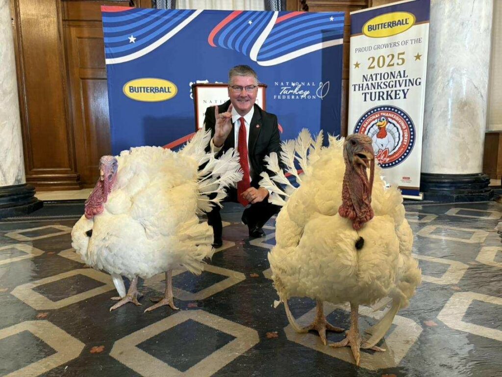 CALS Dean Garey Fox kneels behind Gobble and Waddle, two large white turkeys, at the Willard InterContinental hotel in Washington, D.C., with National Thanksgiving Turkey signage in the background.